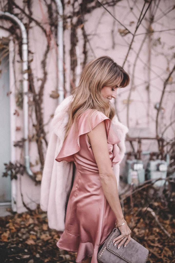 Hottest Pink Holiday Dress featured by top Indianapolis fashion blog, Karina Style Diaries: woman walking in the street wearing a BB Dakota pink ruffle dress, ASOS rose gold sandals, Daniel Wellington watch, grey suede clutch, gold disc necklace and a pink H&M Faux Fur coat